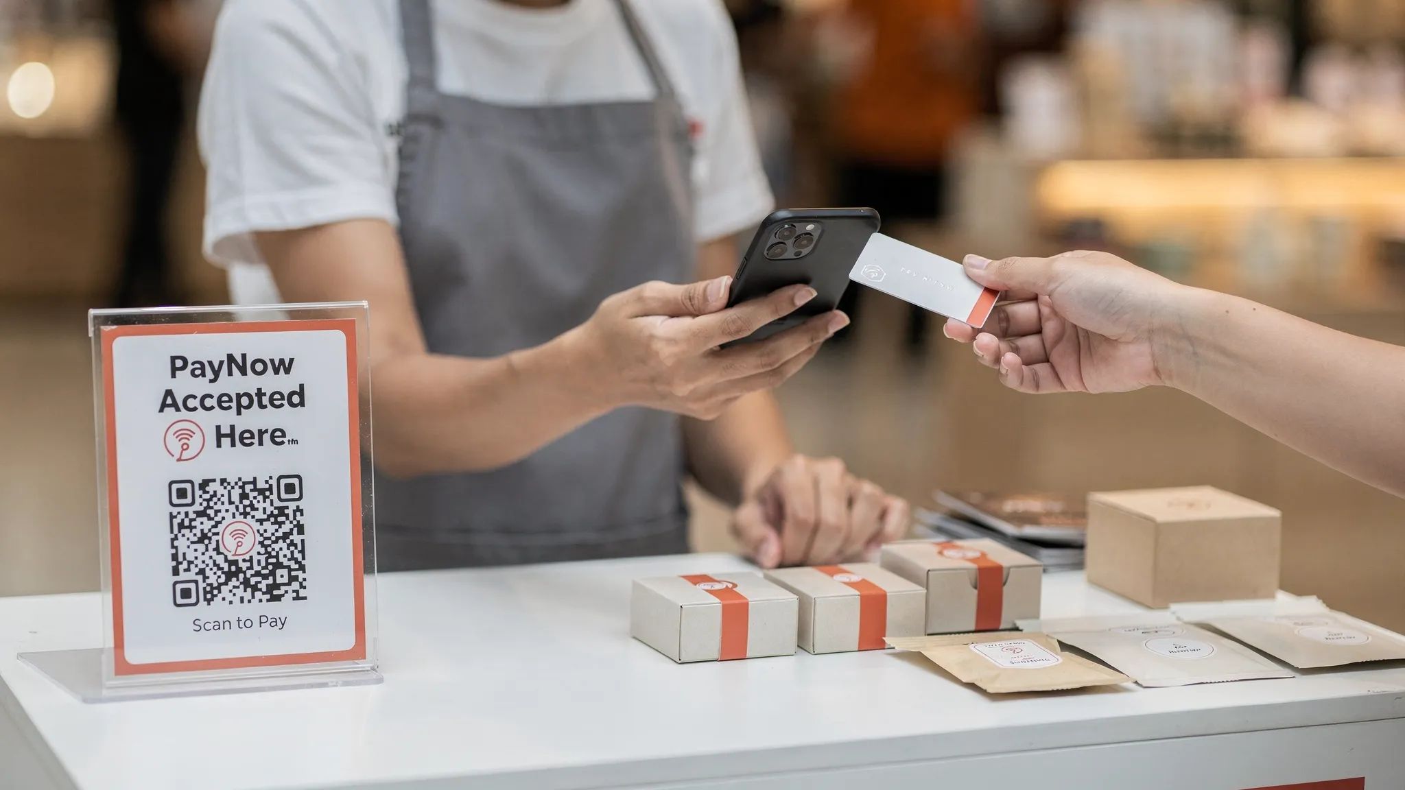 Over-the-shoulder close-up of a Singapore small business owner holding a smartphone as a customer taps a contactless card on the device; a PayNow QR sign sits beside simple product packaging on the pop-up counter.