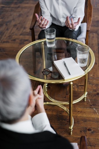 A photo of a two people conversing while sitting on chars with glass table between them