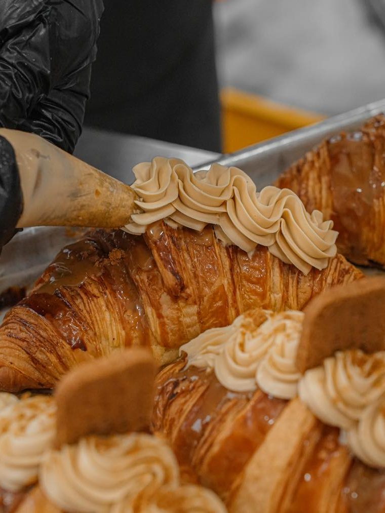 A close-up of a chef piping cream onto a caramel-glazed croissant, adorned with cookie pieces.