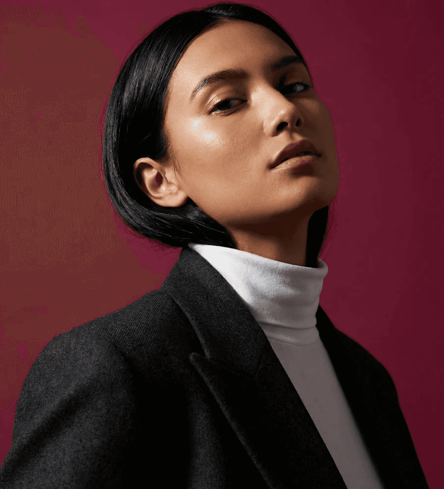Person with short black hair and confident expression, wearing a white turtleneck and dark blazer. The background is a rich maroon color.