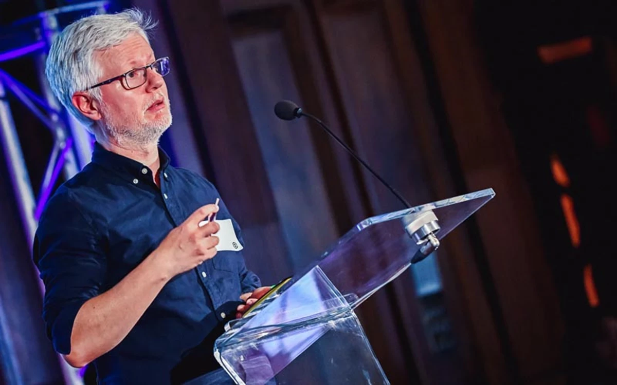 Person standing behind a clear podium with a microphone, holding a small object and speaking on stage with blue and purple lighting in the background.