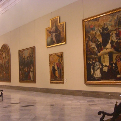 Une galerie avec de grands tableaux classiques exposés sur des murs beiges. Le sol est en marbre et il y a quelques bancs en bois foncé.