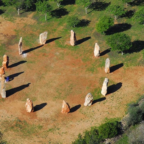 Aerial view of a stone circle monument surrounded by grass and trees, with large standing stones casting shadows.