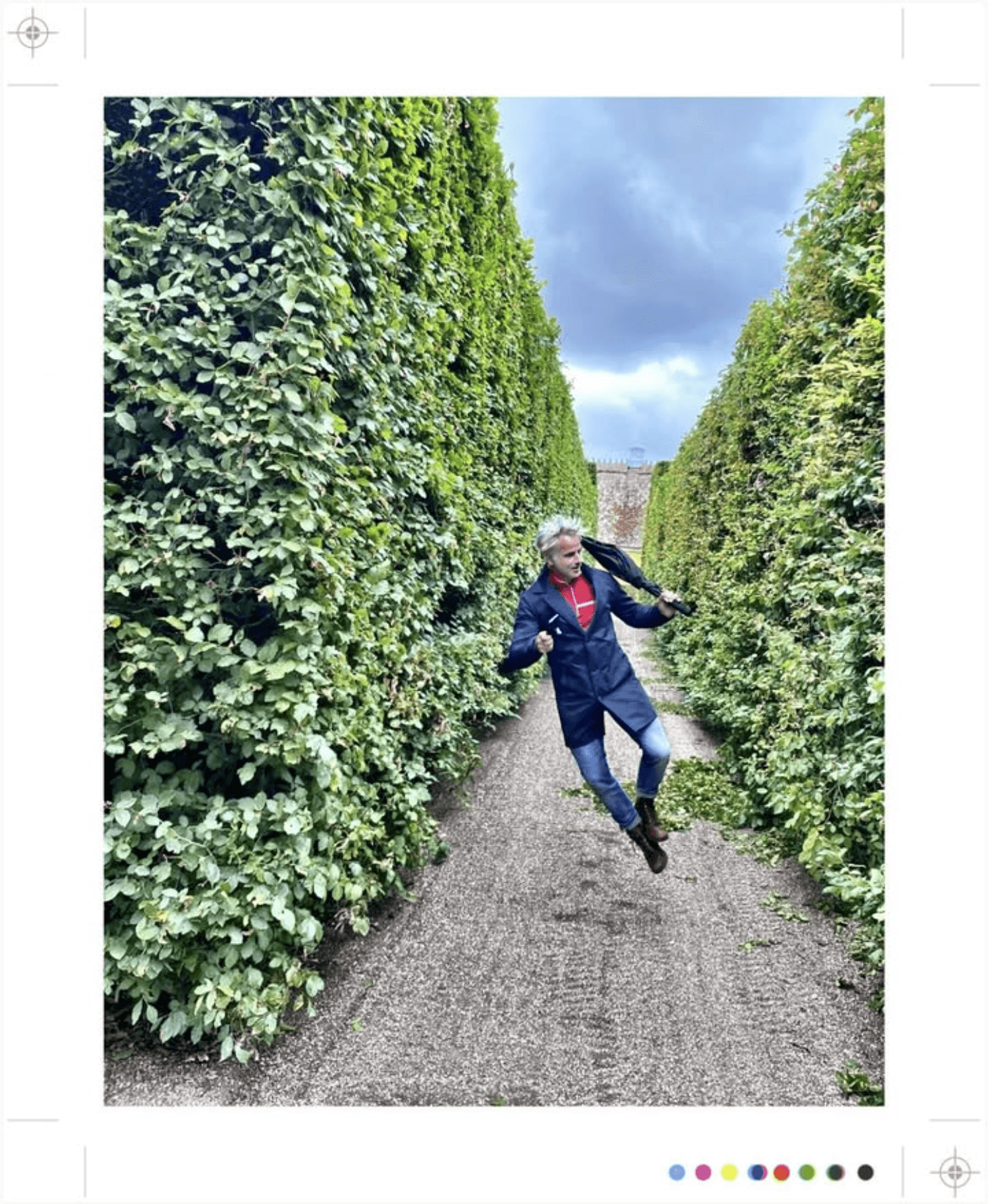 Man in blauwe jas springt in een tuinpad met hoge groene heggen.