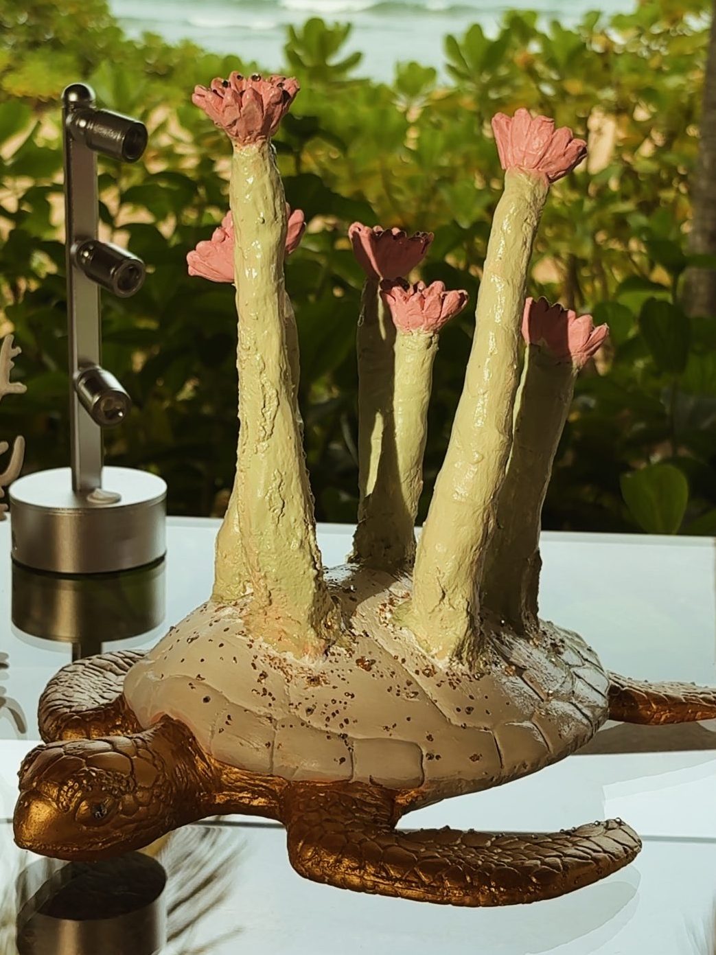 A creatively designed turtle sculpture with flower-like structures on its back, placed on a reflective surface with a beach and ocean in the background.