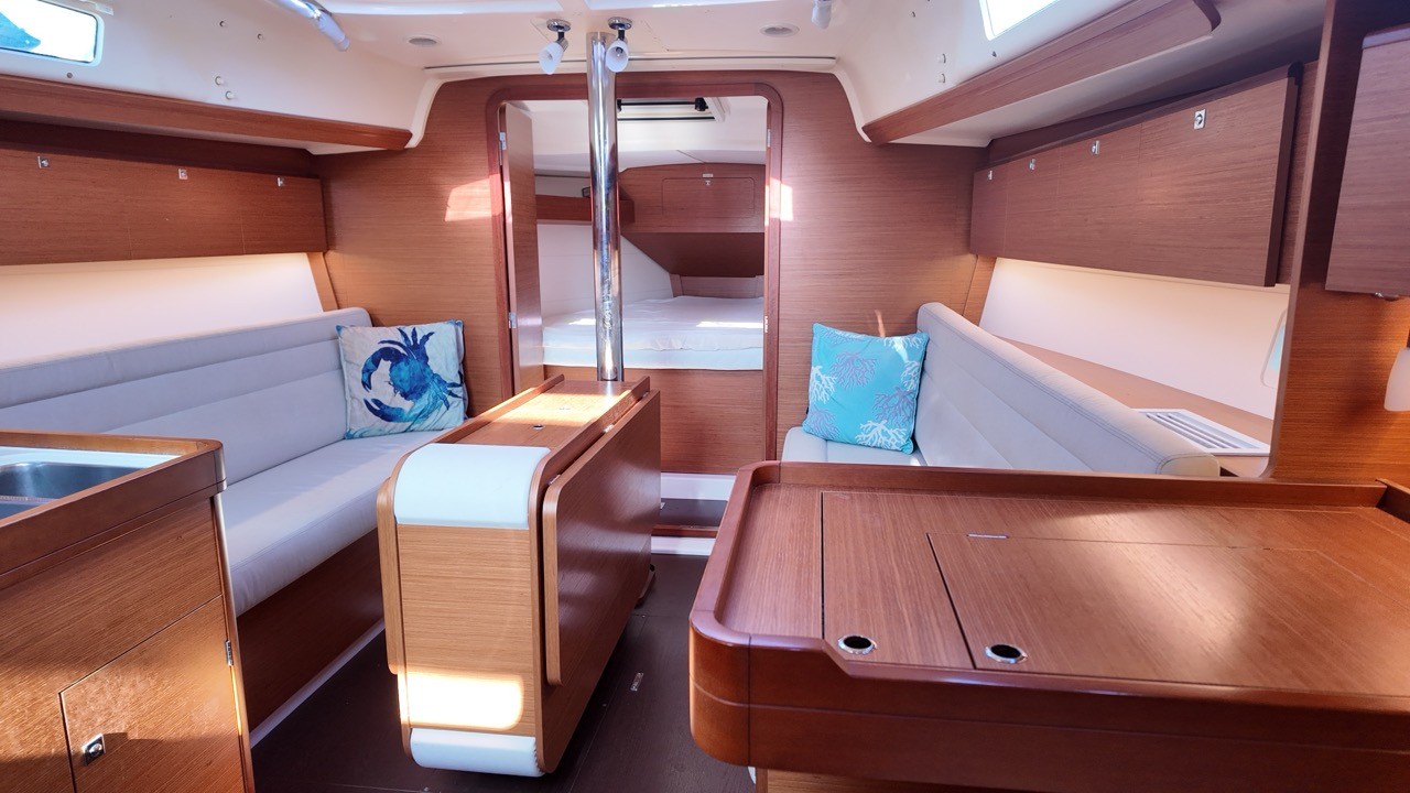 Interior view of the Dufour 350 GL's saloon showing gray settees with blue accent pillows, a fold-out dining table, and a varnished teak chart table with cup holders in the foreground.