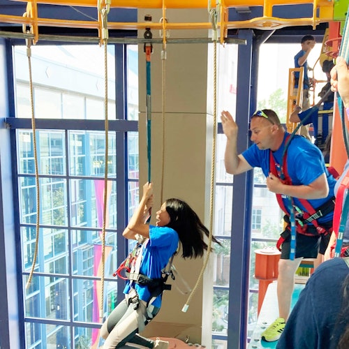 A woman in a harness climbs a ropes course indoors with a guide assisting; large windows are visible in the background.