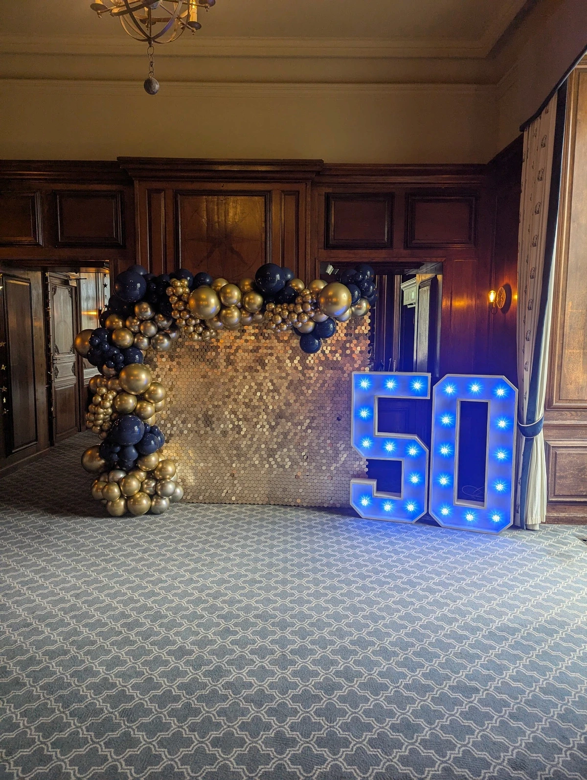 Navy and gold balloon arch around illuminated “50” sign inside a wood-panelled room