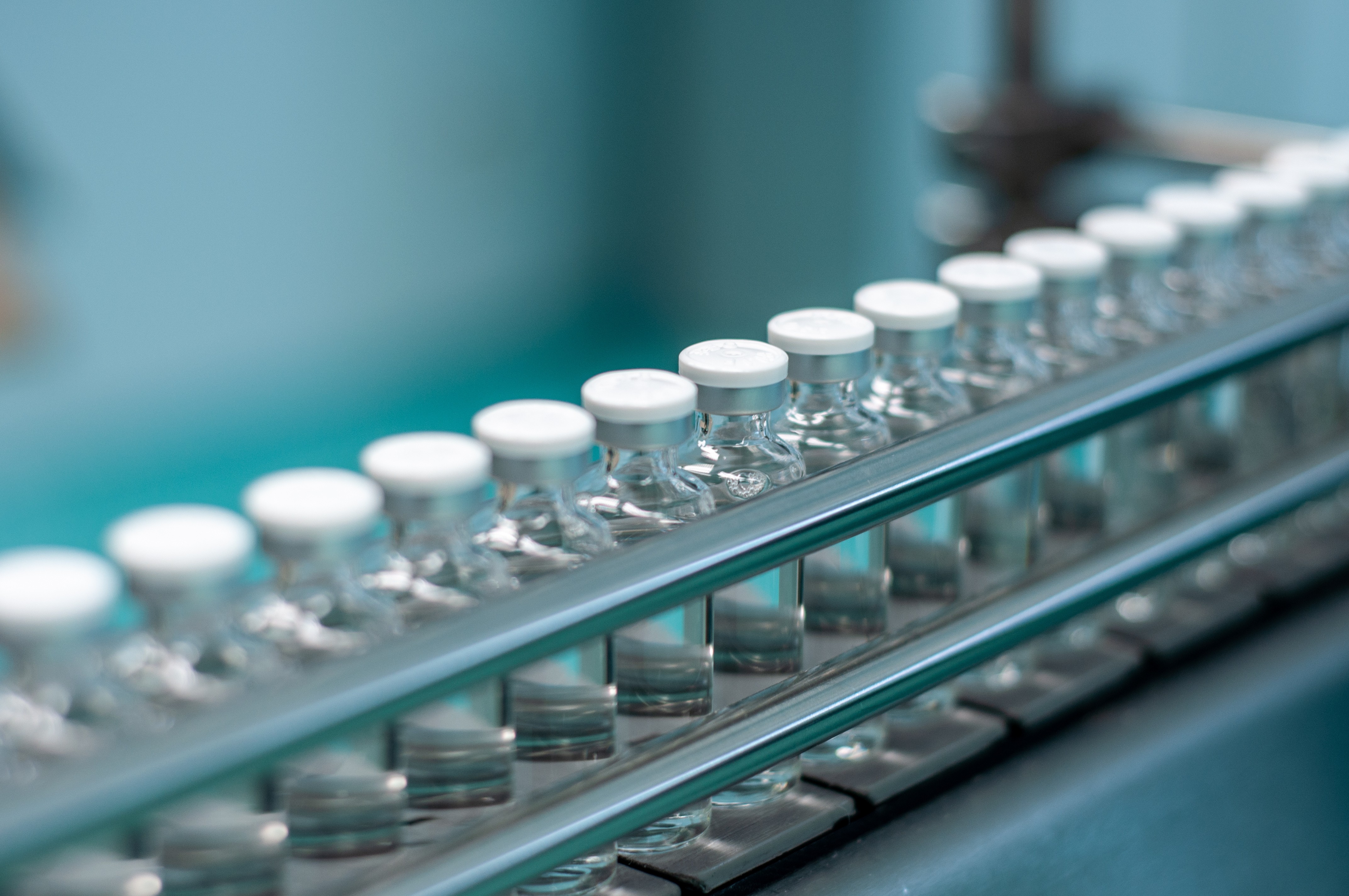 Row of clear capped medicine vials on a stainless steel production line against a teal background