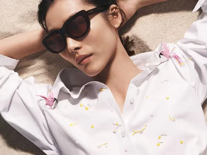 Model in oversized sunglasses and an embroidered white shirt lying on the sand