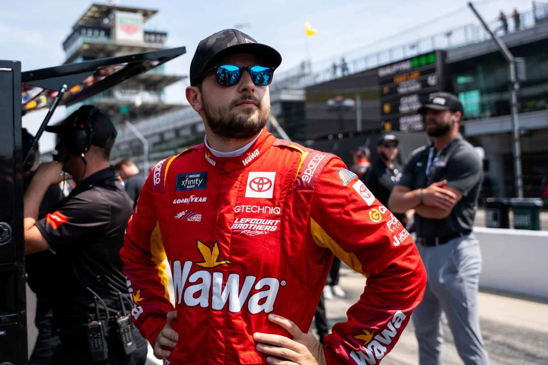 NASCAR driver Joe Graf Jr. wearing a red Wawa racing suit and sunglasses at the track before a race, representing brand sponsorship visibility.