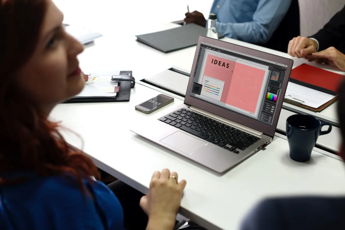 A close-up of a business meeting table, focusing on a laptop displaying design software with the word "IDEAS" prominently on screen. A woman with auburn hair is partially visible in the foreground, suggesting a creative or design discussion is taking place.