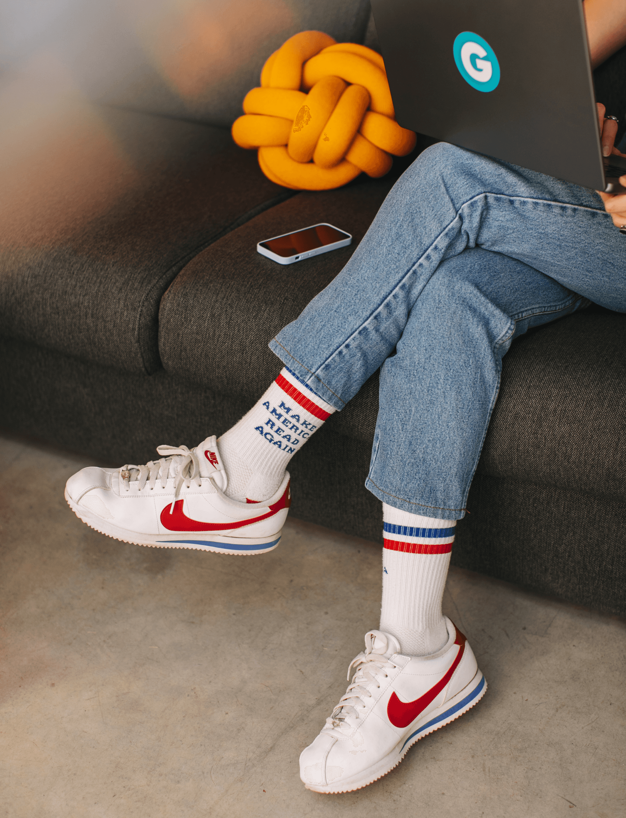 A girl sitting on a sofa with a laptop on her lap, visible from the waist down, wearing Nike sneakers and jeans.