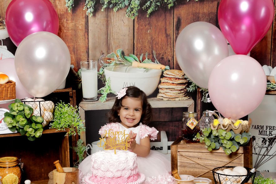 A baby girl with a pink dress is sitting in a rustic cake smash session with a wooden backdrop, pink-and-white balloons, and a pink birthday cake.&nbsp;