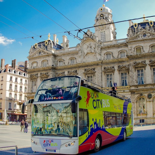 A colorful double-decker Lyon City Bus in front of an ornate historic building under a clear blue sky.