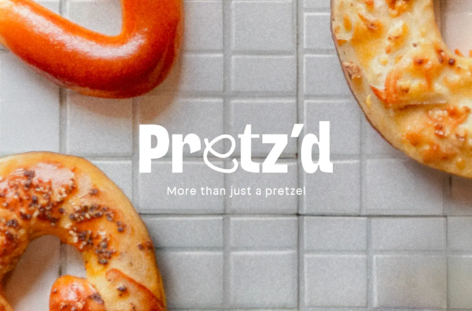 Vue aérienne de plusieurs bretzels artisanaux sur carrelage gris avec le logo Pretz'd et la baseline "More than just a pretzel" en blanc.