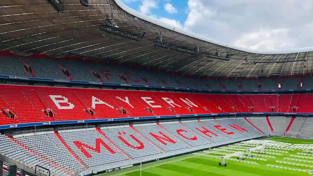 Die Allianz Arena in München, das Heimstadion des FC Bayern, ist leer und ruhig, mit Sitzen, die das Logo ‚FC Bayern München‘ zeigen. Dieses Stadion ist die Arbeitsumgebung von Max Eberl, dem neuen Sportdirektor des FC Bayern, der seine umfassende Erfahrung im Fußballmanagement nutzt, um den Verein durch eine Phase der Transformation und des Erfolgs zu führen.