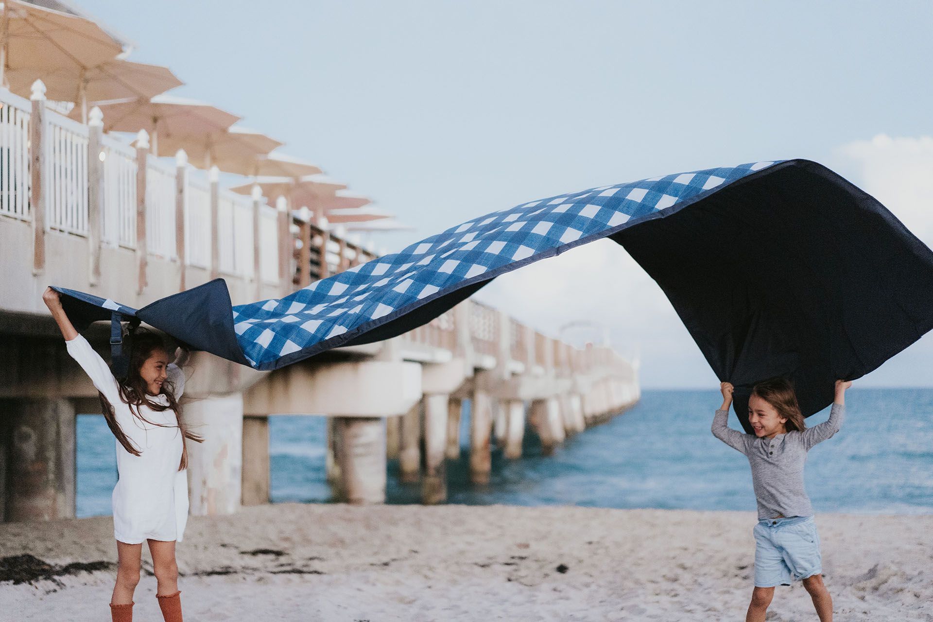 2 kids on the beach waving a picnic blanket