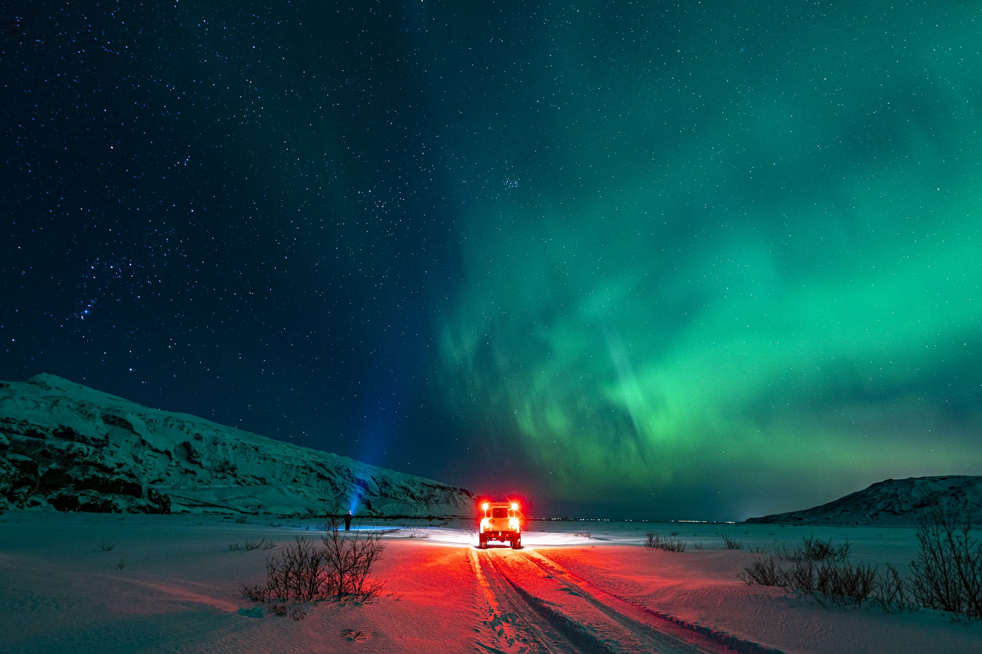 4x4 vehicle parked beneath the northern lights in Iceland with snowy mountains and a star-filled sky.