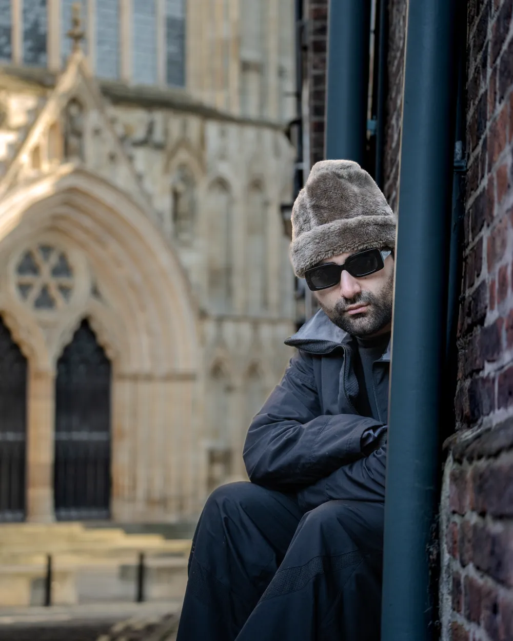 Aria wearing sunglasses and a faux-fur hat, seated in a shadowed corner of Precentor's Court with the blurred stonework of York Minster in the background.