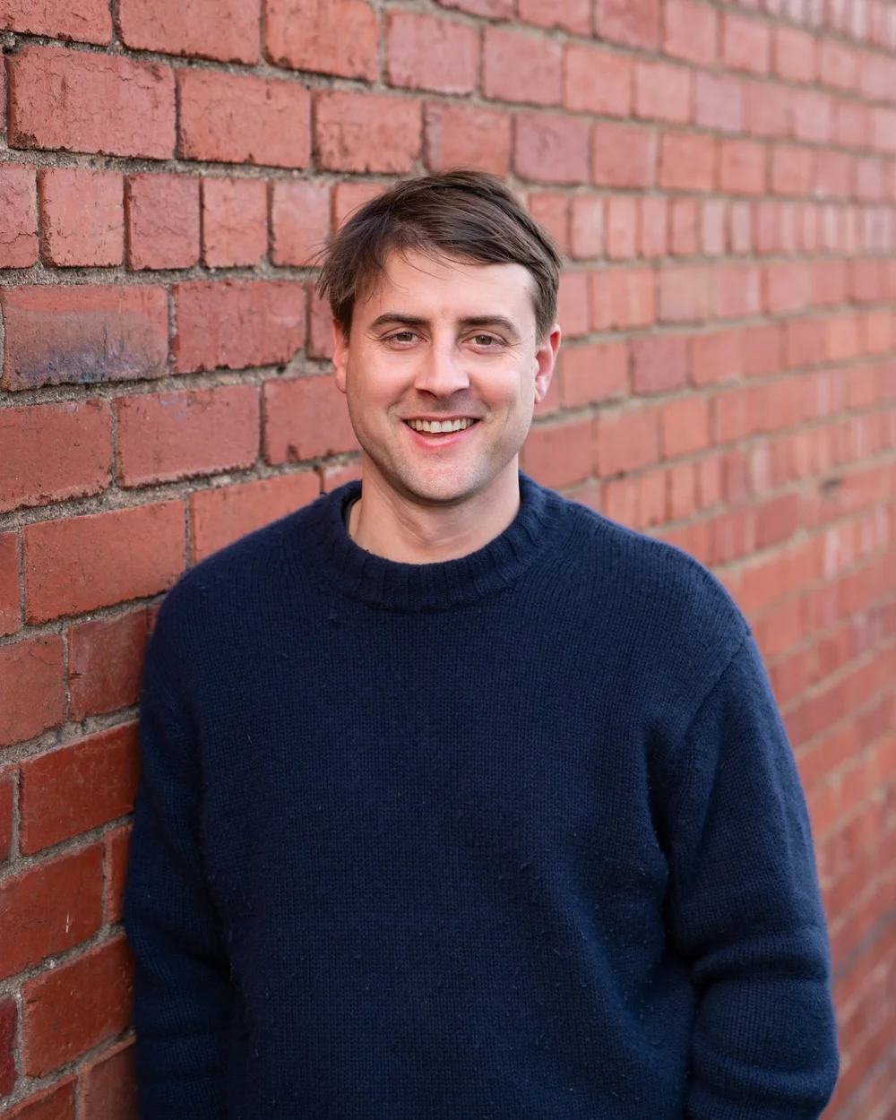 Smiling man in a navy sweater standing against a red brick wall.