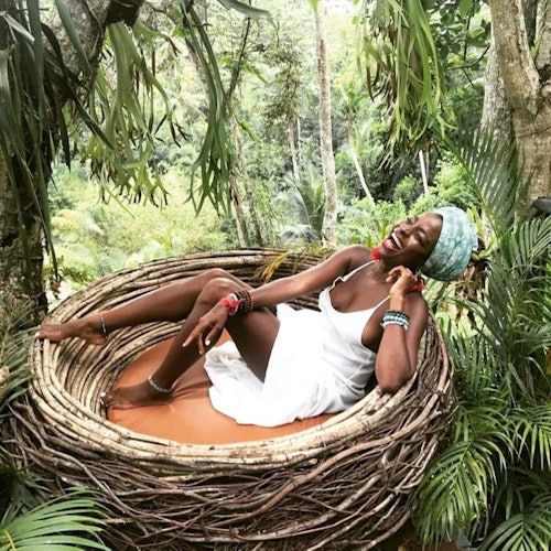 A person in a white dress relaxes in a large, nest-like structure surrounded by lush greenery, smiling joyfully.