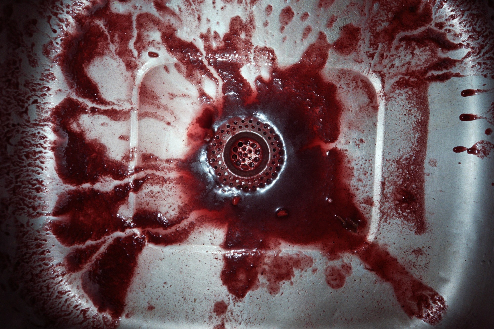 A stainless steel sink stained with dark red liquid splatters around the drain, resembling spilled wine or juice, creating a striking contrast against the metal surface.