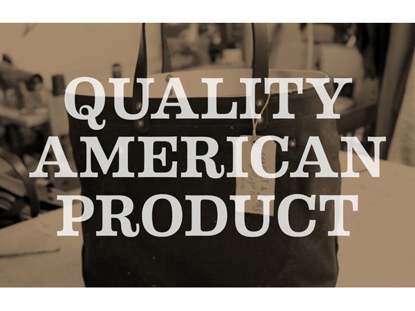 A black tote bag with two dark handles sits on a light-colored surface. The words “QUALITY AMERICAN PRODUCT” are superimposed in white, bold, sans-serif font across the center of the image. The background is out of focus and appears to show a workspace with indistinct shapes and objects.