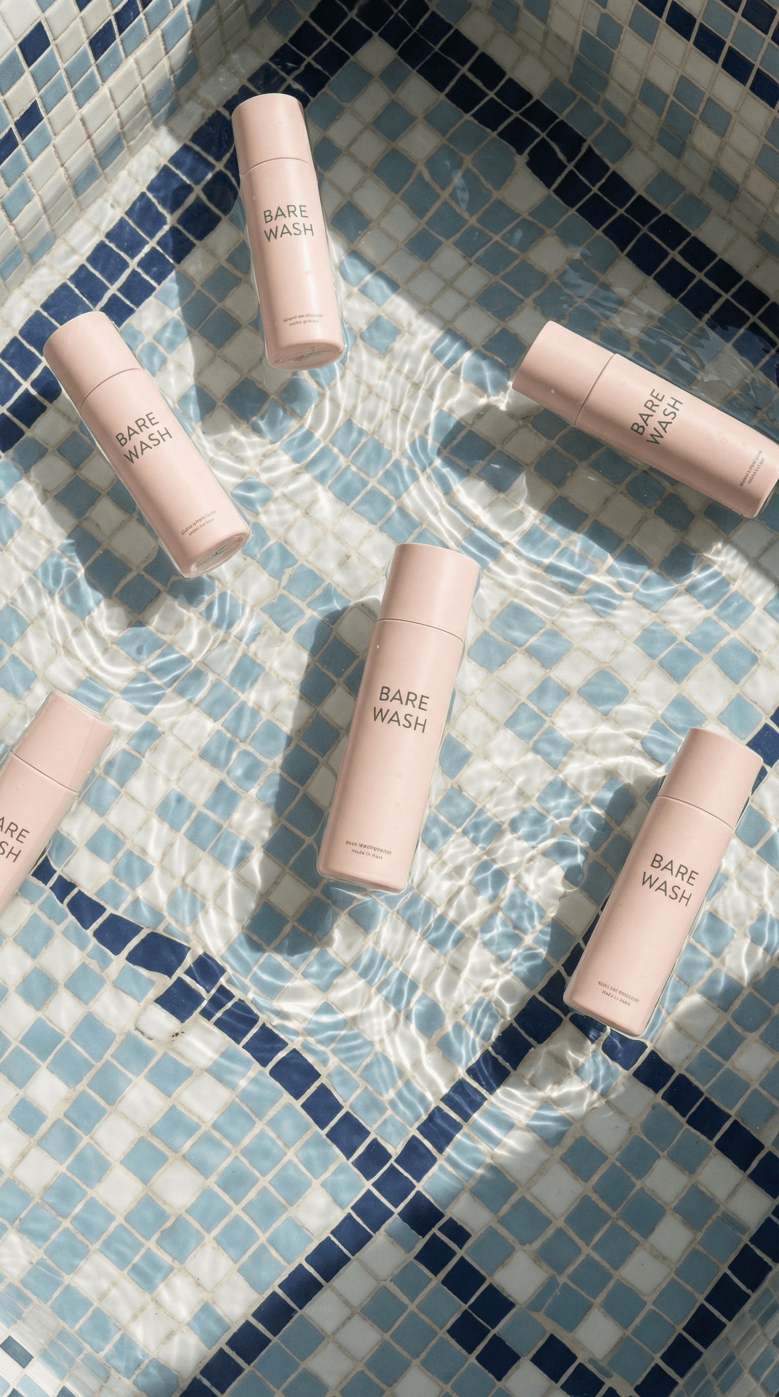 Bare Wash body cleanser bottles floating in blue mosaic pool water with sun reflections