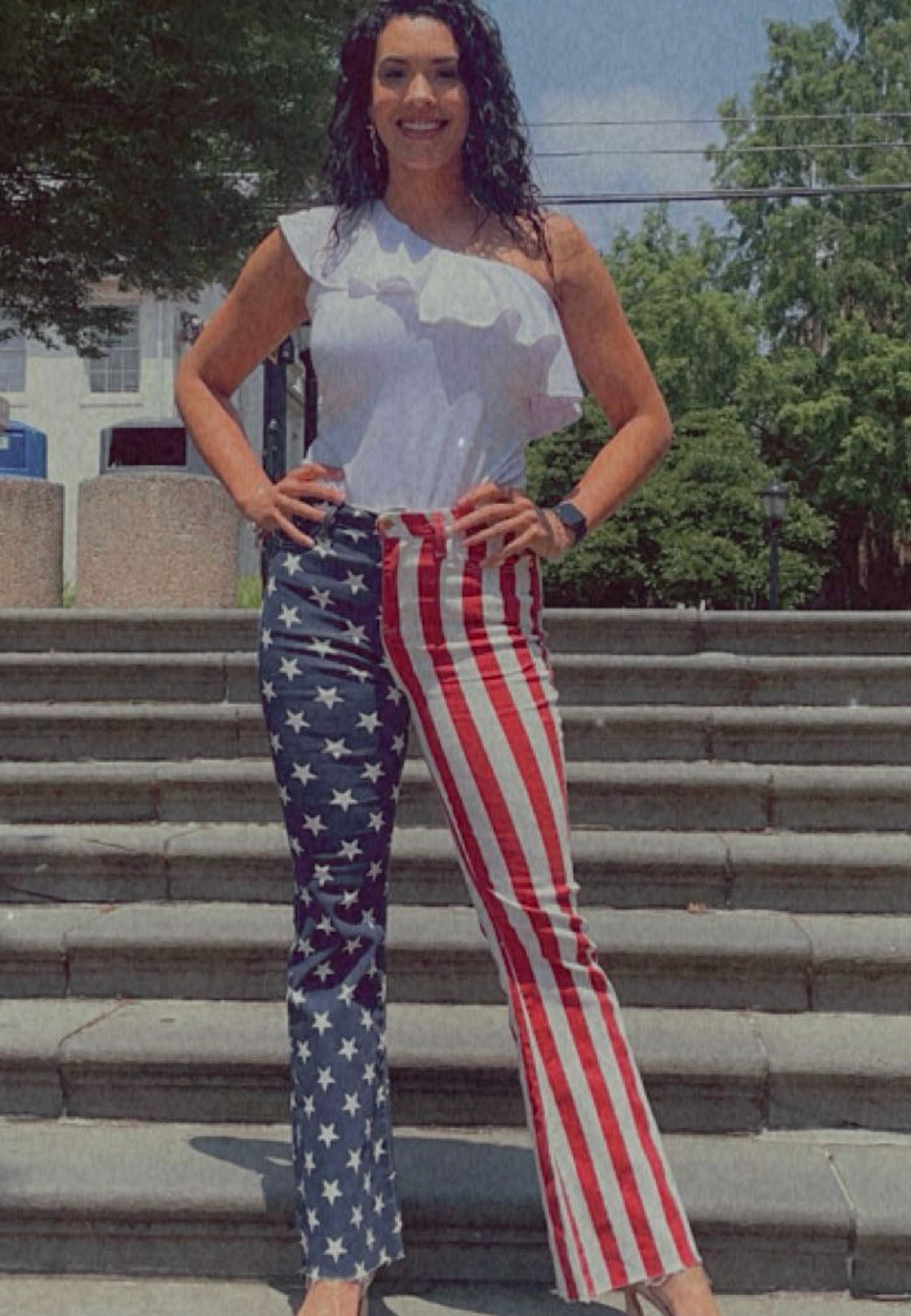 A woman stands confidently on outdoor steps, wearing a white ruffled top and American flag-themed pants. The background features greenery and buildings.