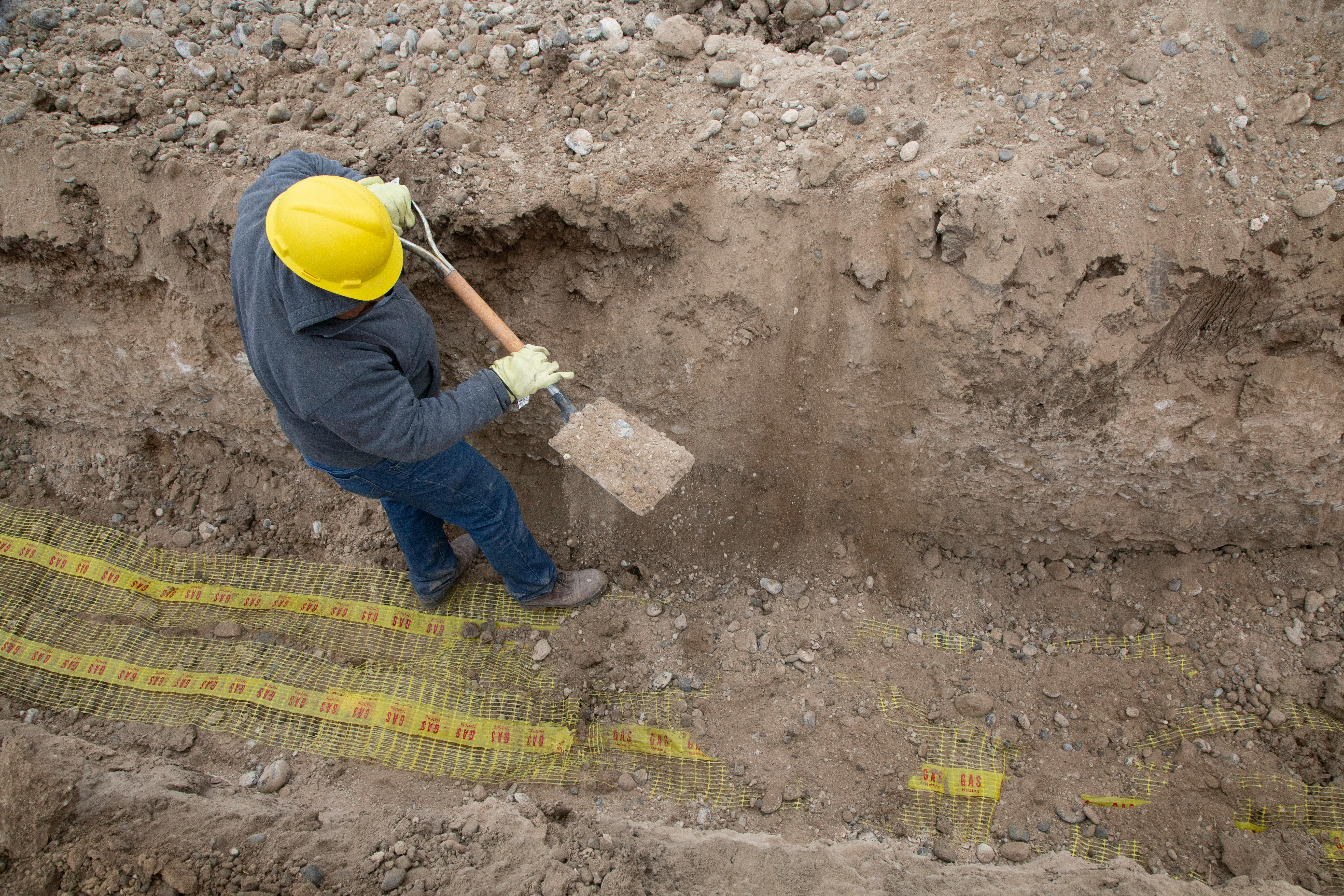 Worker in a hard hat digging a trench with a shovel.