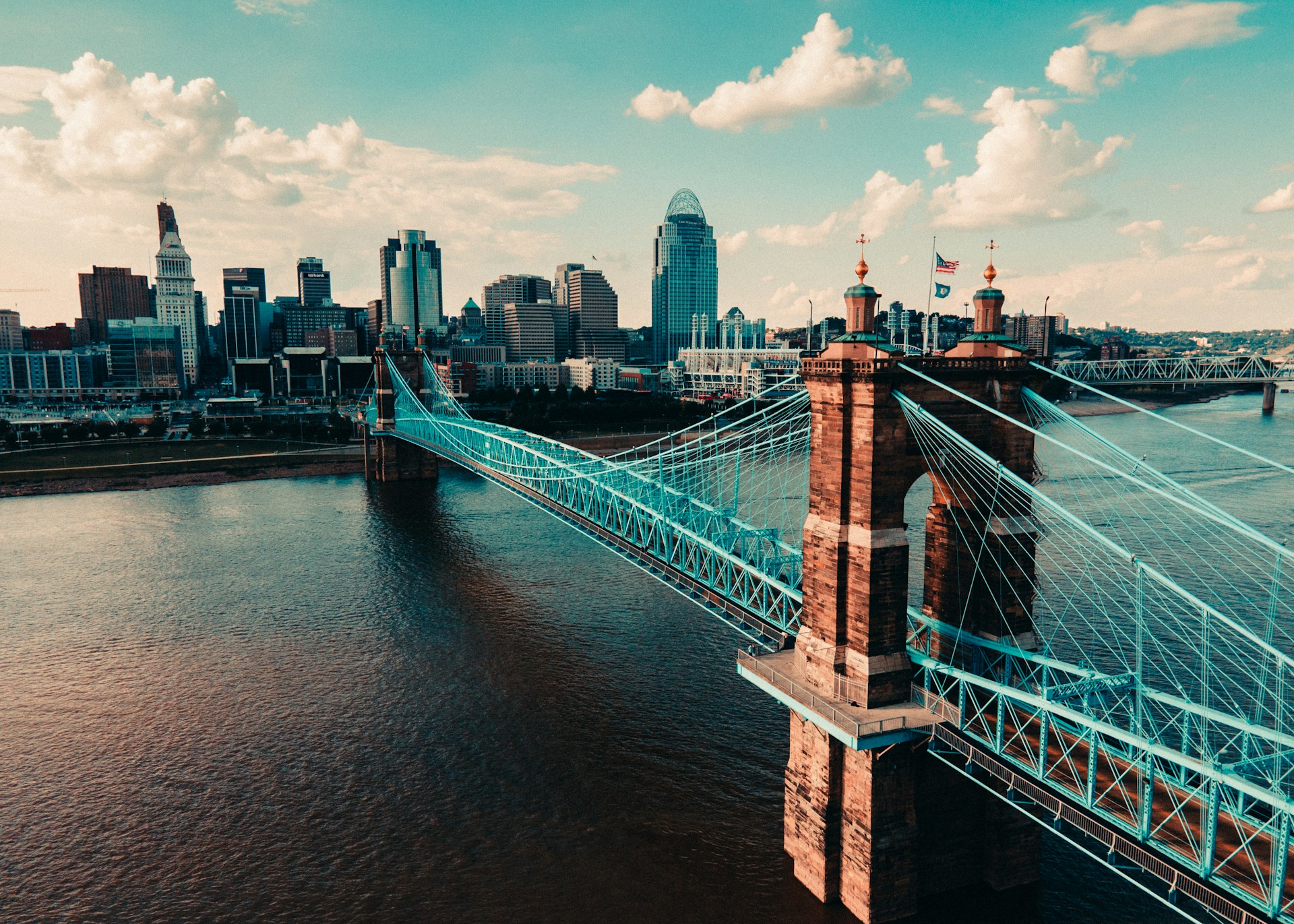 Suspension bridge with tall brick towers spanning a wide river with a city skyline behind.