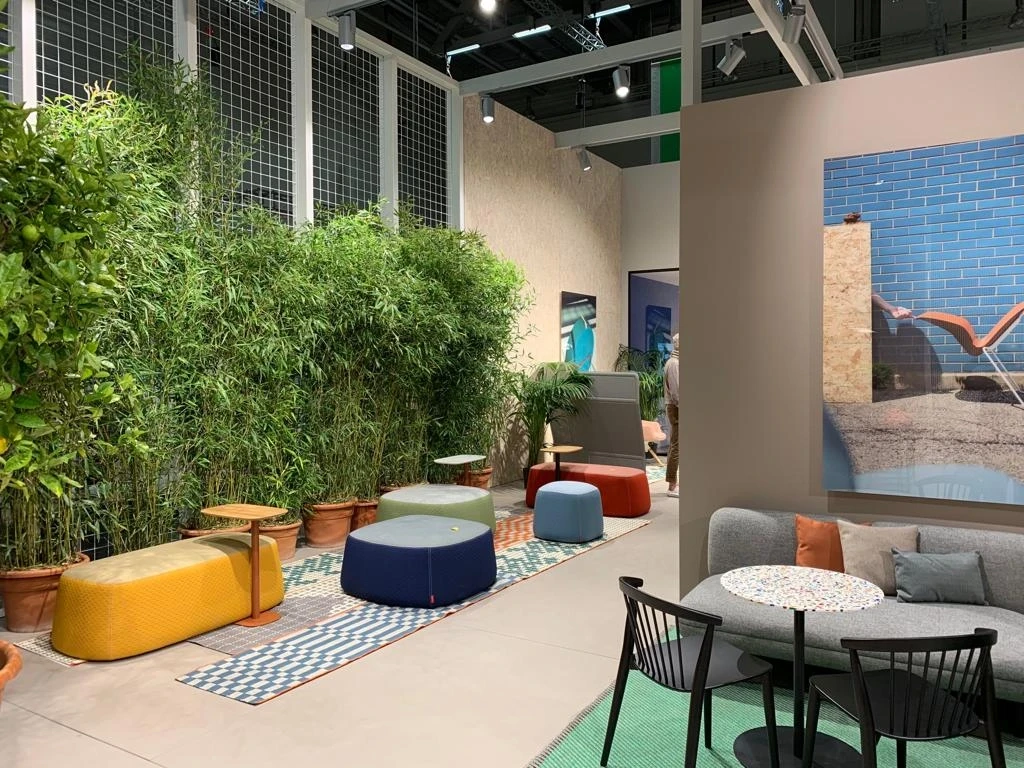 Garden-style lounge at the HAWORTH ORGATEC stand with bamboo plants in terracotta pots, colorful poufs on patterned rugs and a grey sofa with terrazzo table.