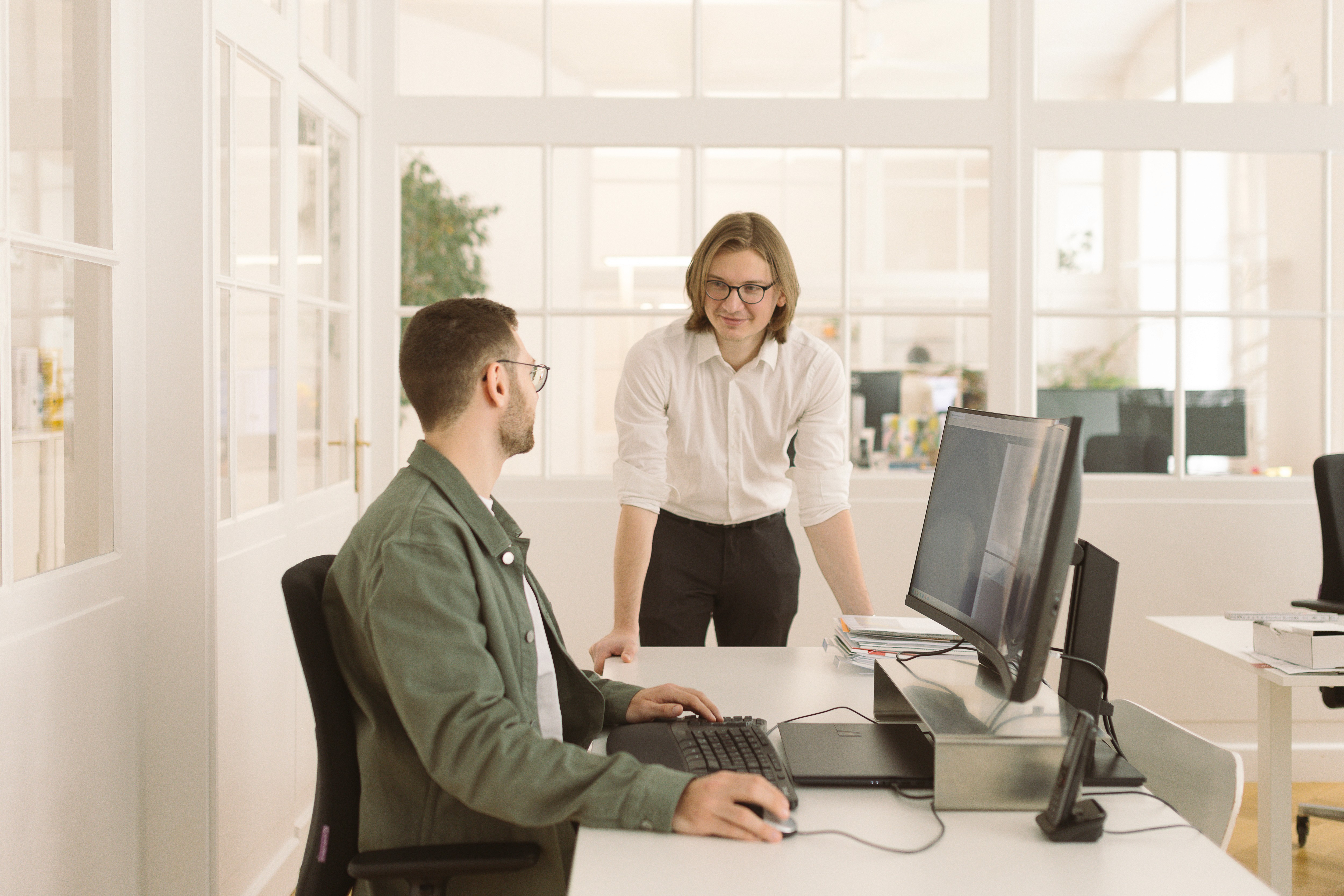 Two male colleagues having a conversation in a bright, modern office. One person sits at a desk working on a computer, while the other stands, leaning casually on the desk and smiling. The atmosphere is friendly and collaborative.