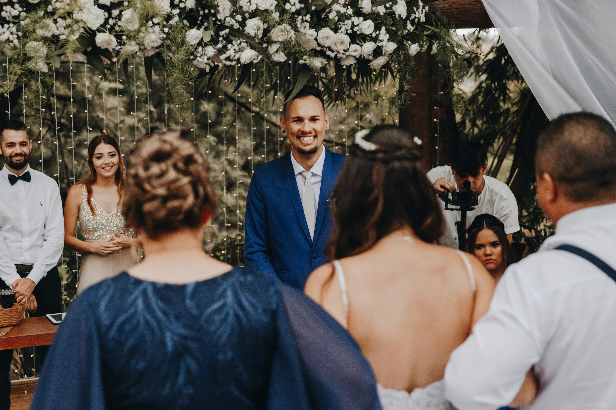 Groom smiling at the altar under a white floral arch with string lights as the bride walks down the aisle with her family