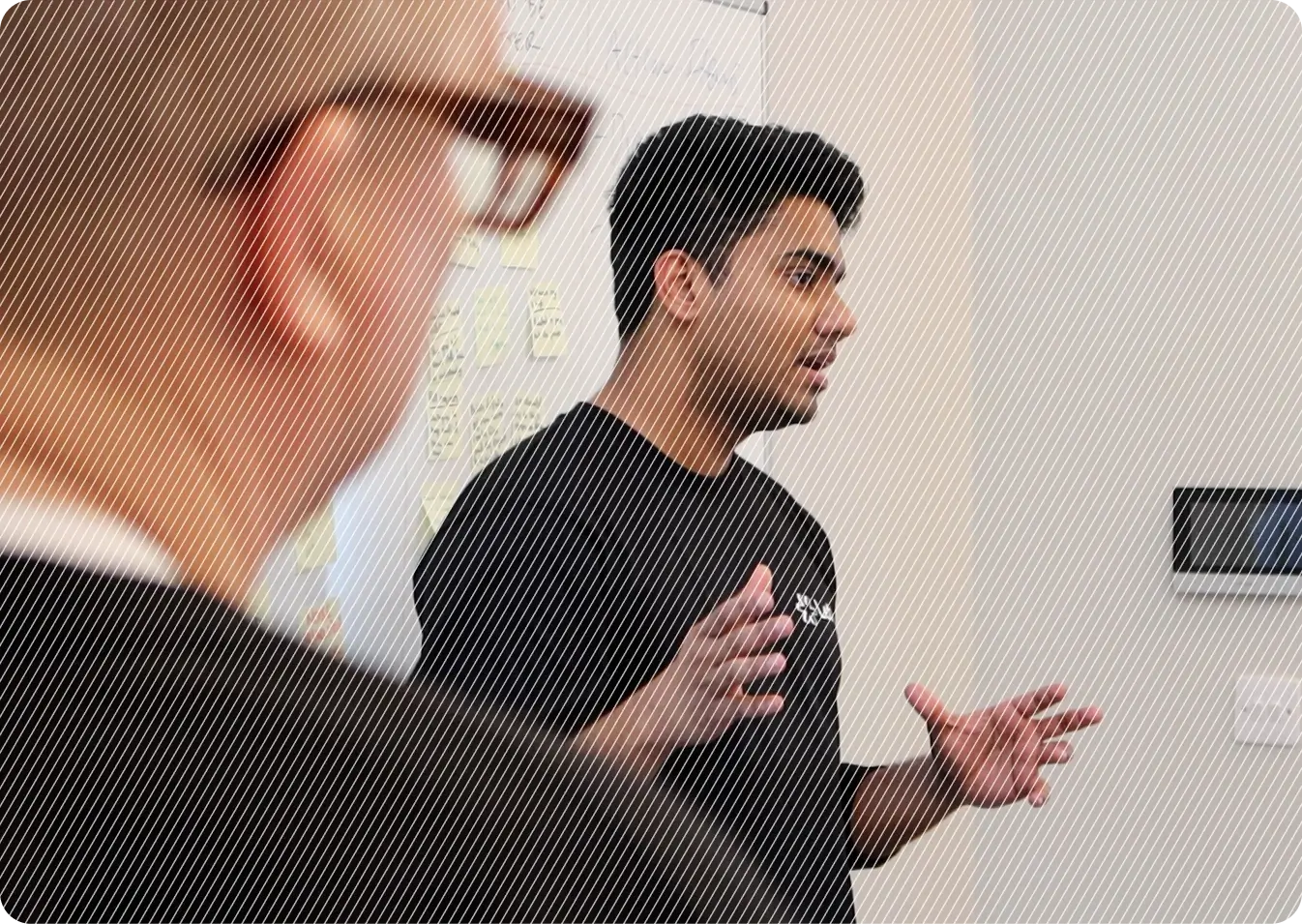A person is speaking and gesturing during a meeting, with a whiteboard covered in sticky notes in the background and another person in the foreground, slightly out of focus.