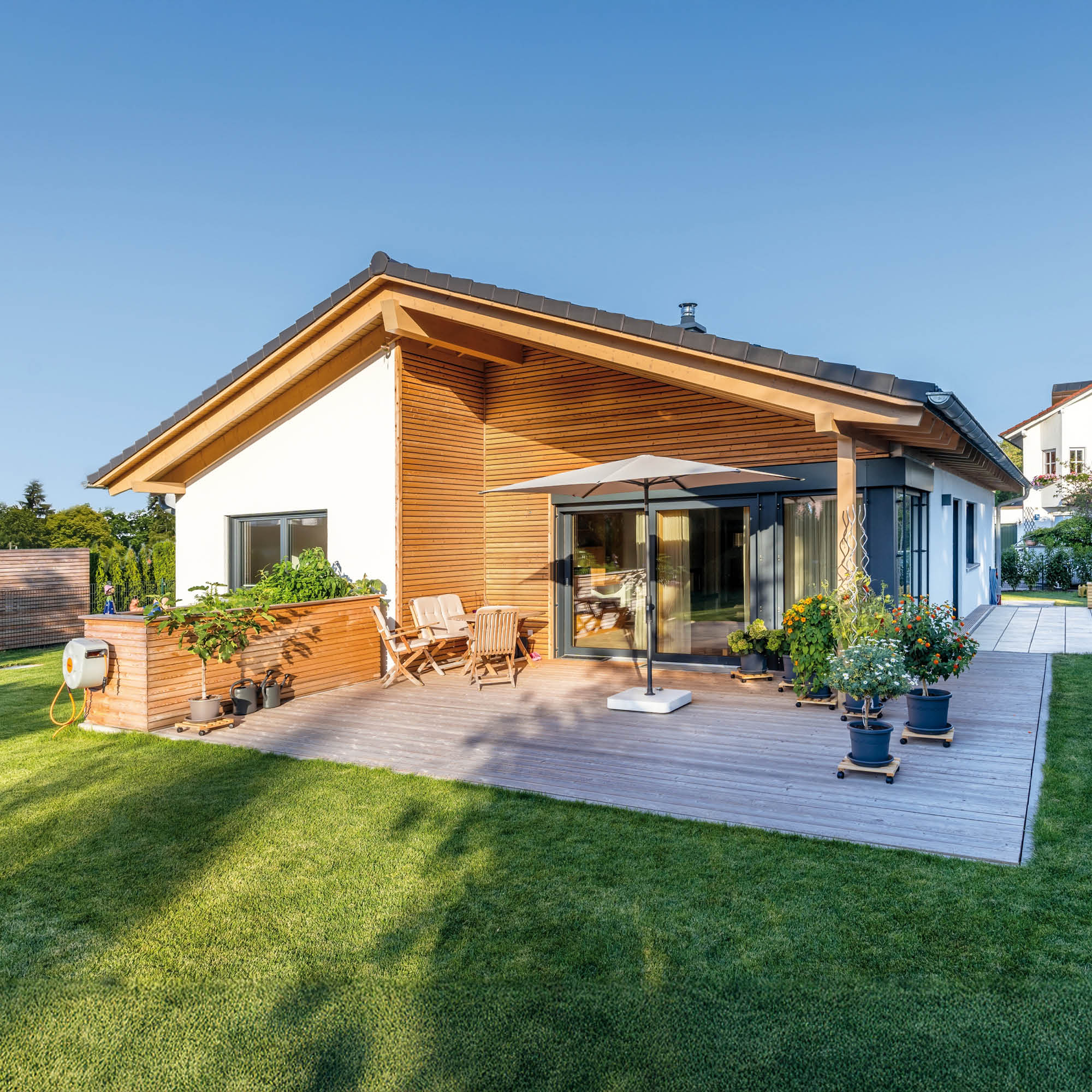 Modernes Einfamilienhaus mit Holzfassade, großer Terrasse mit Möbeln, Sonnenschirm und Garten.