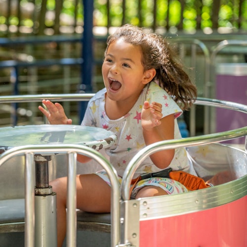 Seorang gadis muda dengan gembira menaiki permainan berpusing di taman hiburan, sambil tersenyum dan bertepuk tangan.
