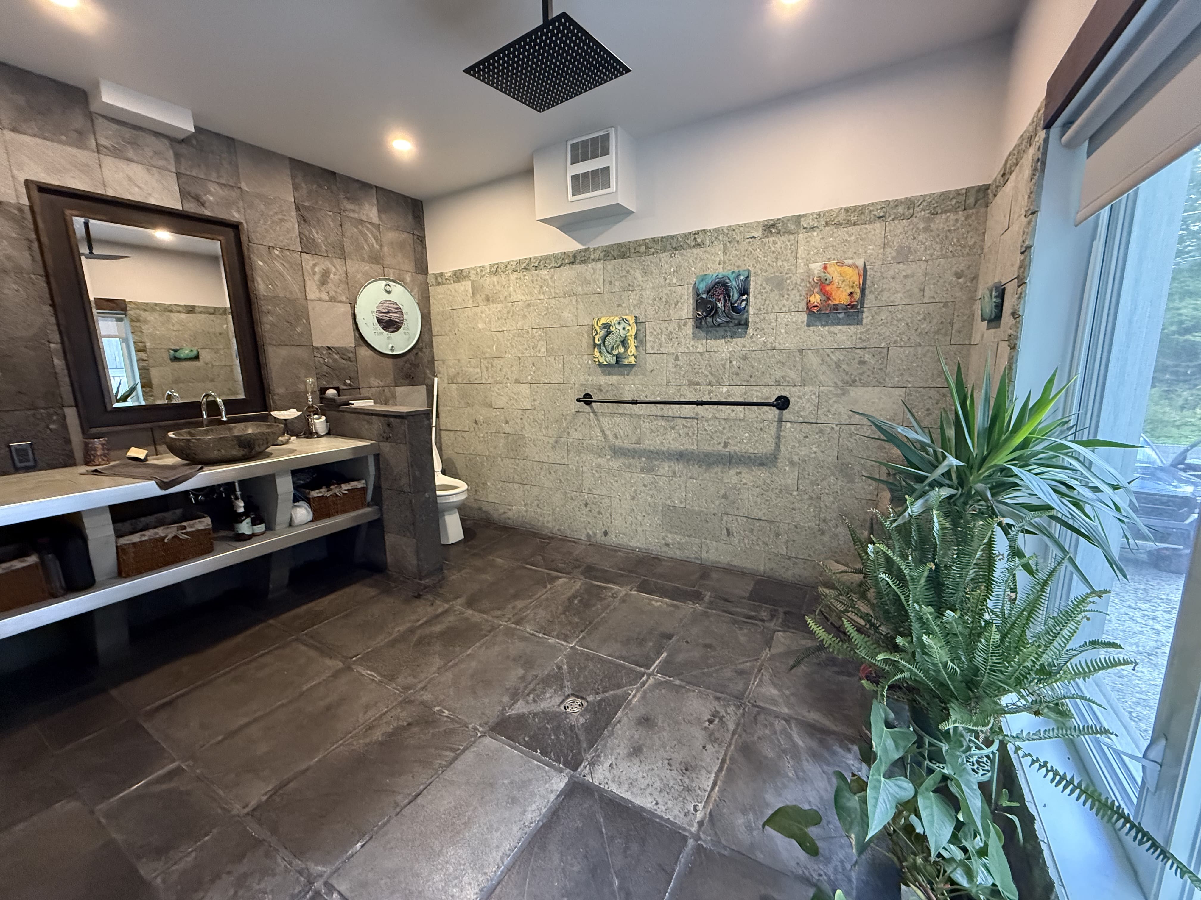 Grande douche italienne avec pommeau pluie, murs en pierre grise et plantes vertes, ambiance zen et naturelle.