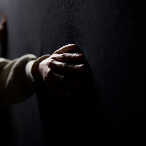 A hand in a white sleeve rests on a dark wall, illuminated by soft light in a low-lit environment.