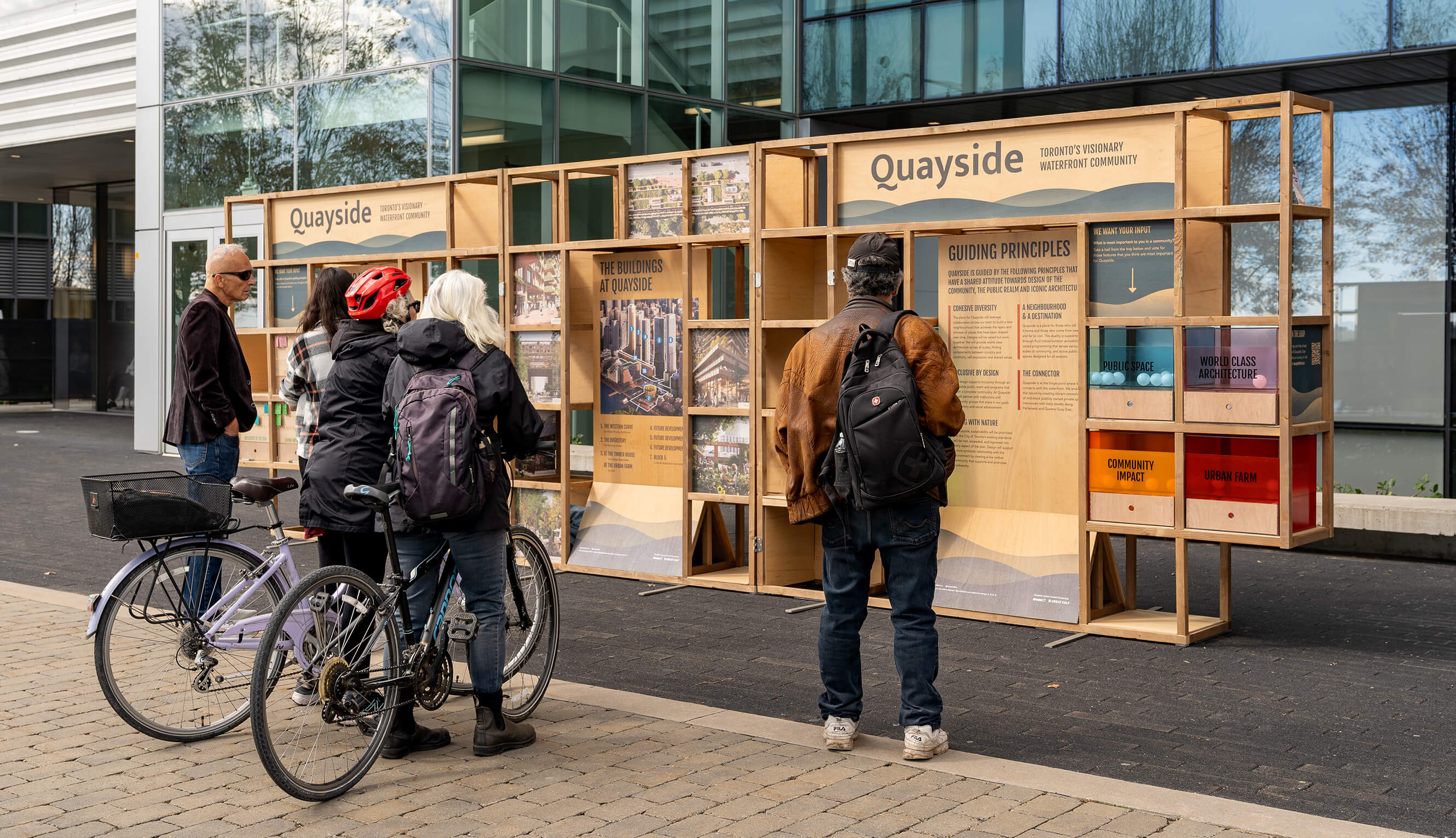 Pedestrian and cyclist visitors interacting with the exhibit strucutre: a large, wooden armature that supports information and architects' renderings printed onto plywood boards, alongside a number of interactive components.