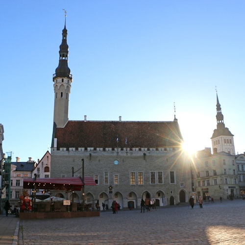 A város főtere történelmi épületekkel, köztük egy nagy épület magas toronnyal, miközben pár ember sétál a tiszta, napos ég alatt.
