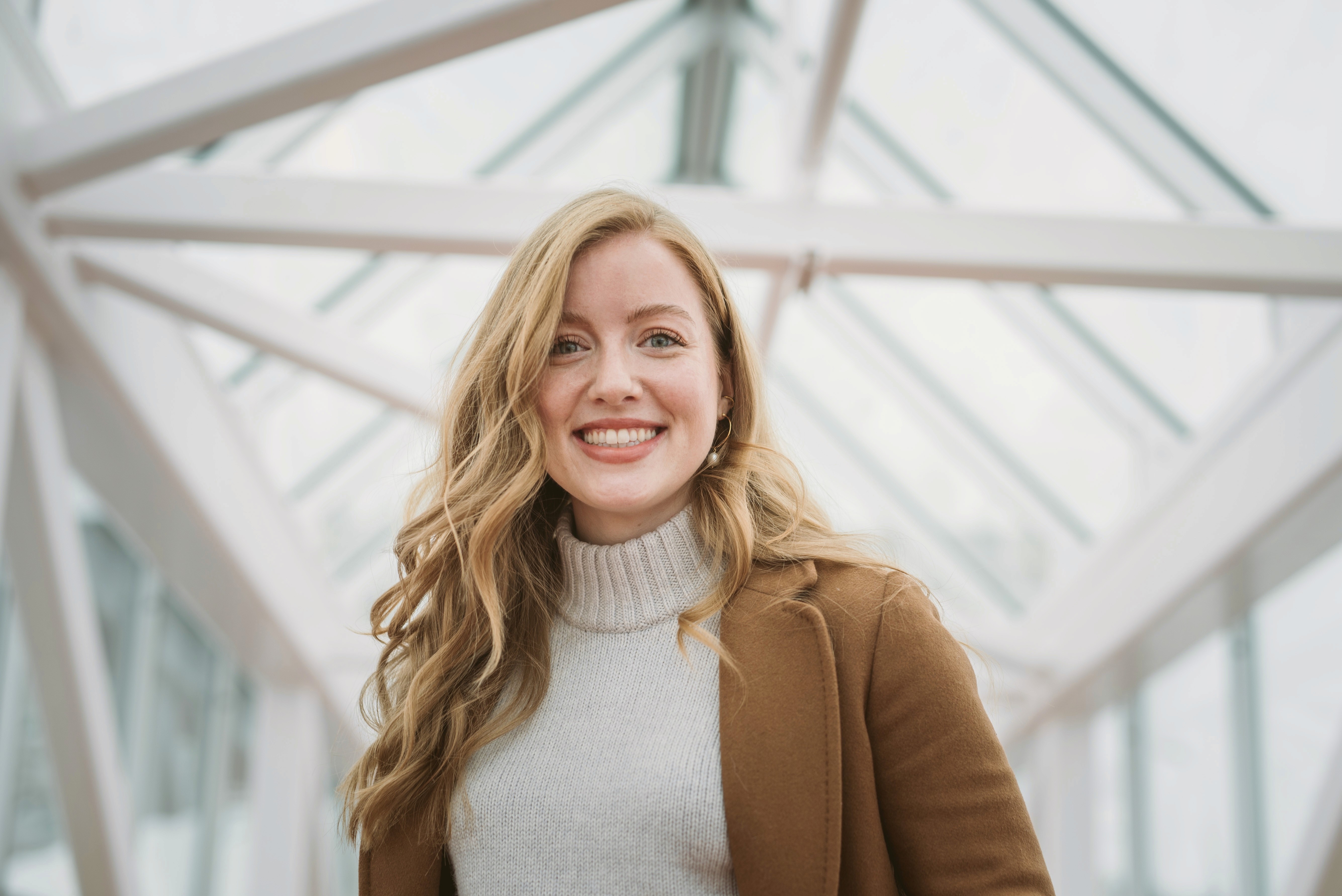 A smiling woman with long wavy hair, wearing a light sweater and a brown coat, stands in a bright indoor setting.