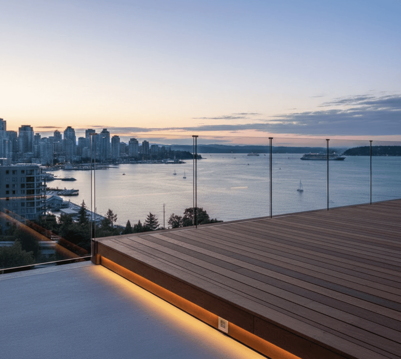 Side angle view of frameless glass railing on elevated waterfront deck, showing mounting hardware and engineering details, English Bay in background, professional construction photography, clean modern aesthetic