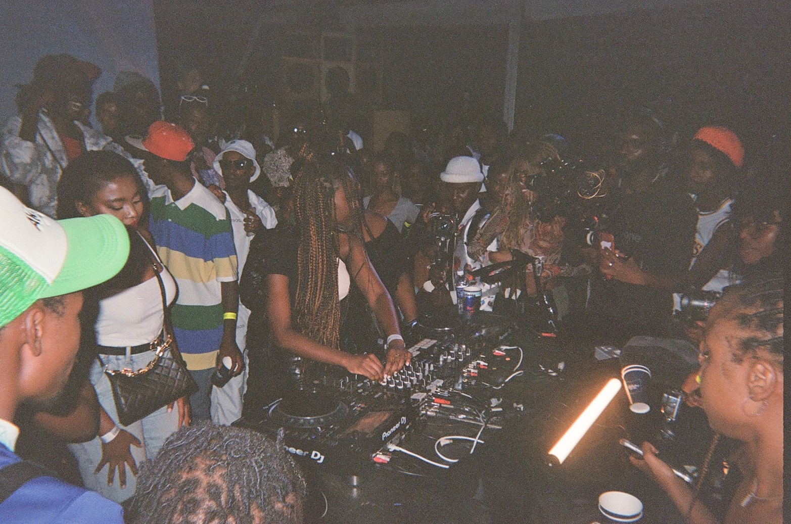 A grainy film photo of DJ Coco Em playing a set while surrounded by people.