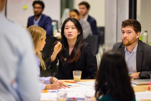 A young person with curly hair smiles at the camera while sitting in front of a laptop against a dark background.