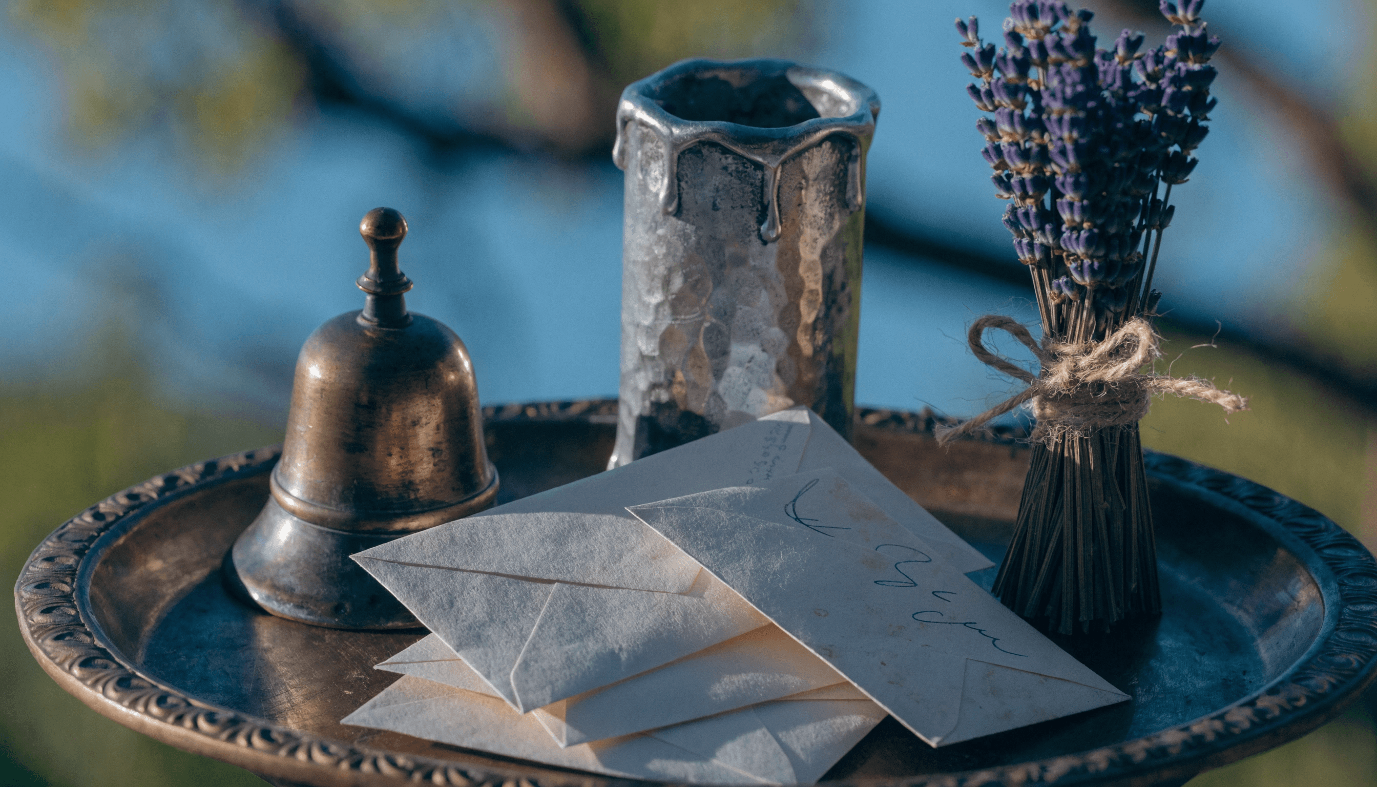 A realistic macro photograph of an #8C7D64 Aged Bronze tray holding several handwritten #F3F6FA Off-White envelopes, a small bronze bell, and a thick #A3AEC4 Sacred Silver candle. A small bundle of dried #7668A6 Magical Lavender is tied with twine. Warm, golden hour lighting with dramatic chiaroscuro shadows.