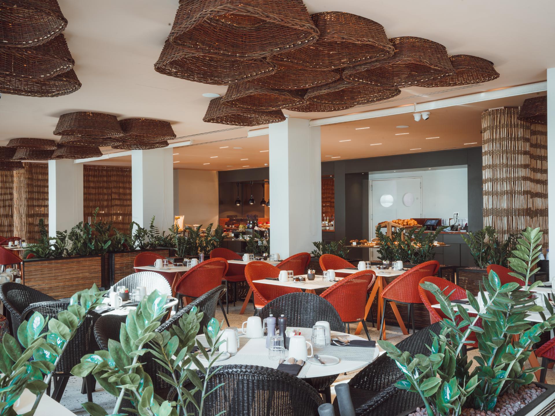 Restaurant mit Tischen, Stühlen und dekorativen Deckenlampen. Pflanzen und gedeckte Tische im Vordergrund.