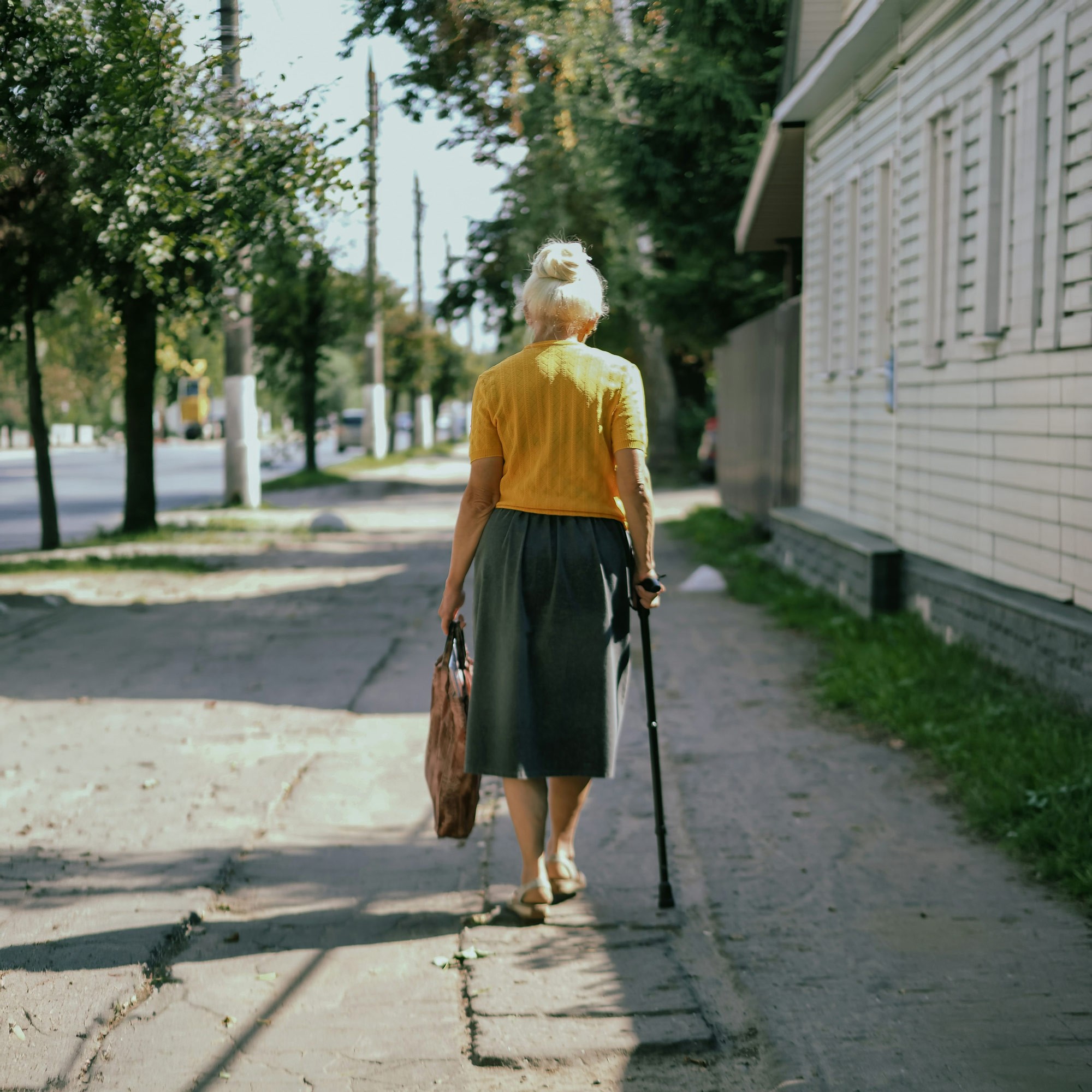 Ältere Frau von hinten, die mit Gehstock und Tasche einen sonnigen Gehweg entlangläuft
