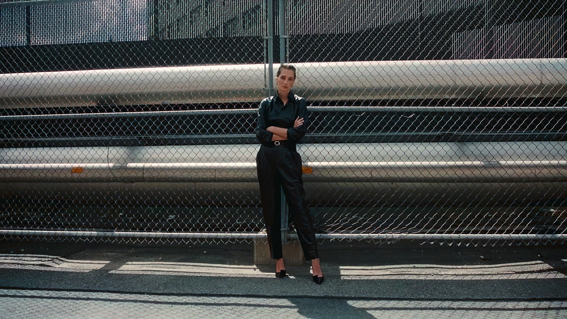 A woman in a black jumpsuit in front of a chain-link fence and large steel pipes, Lafayette 148 New York